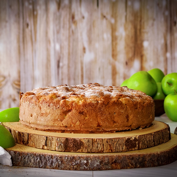 An Irish apple Cake on a wooden cake stand surrounded by green apples.